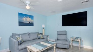 Un salon avec un mur bleu clair, un ventilateur de plafond, un canapé, un fauteuil, une table basse et un téléviseur fixé au mur.