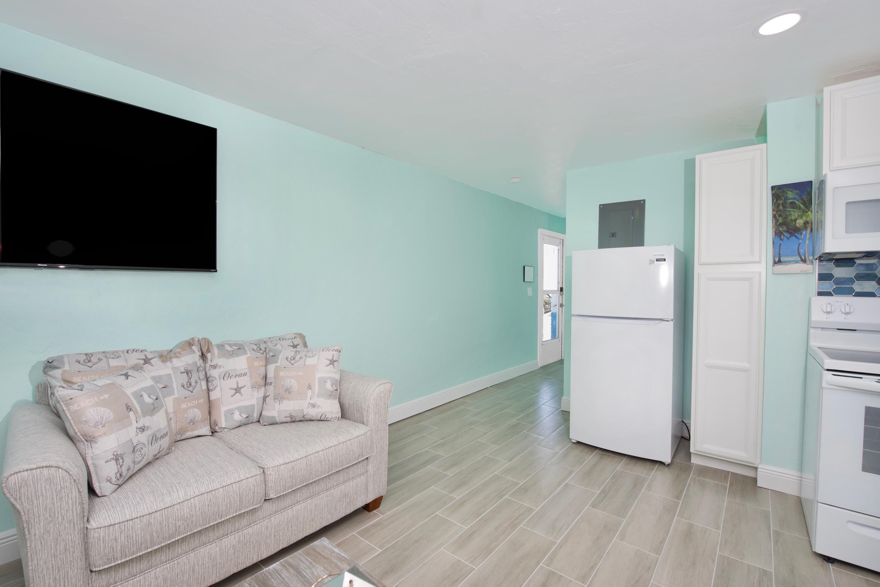 A living room with a light-colored sofa, a flat-screen TV, and a kitchen area with white appliances.