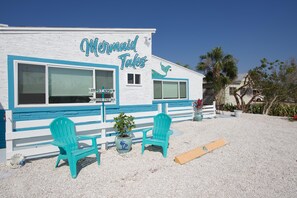 Un bâtiment sur lequel est apposée une enseigne indiquant « Mermaid Tales », avec vue sur la plage, deux chaises turquoise et une plante.