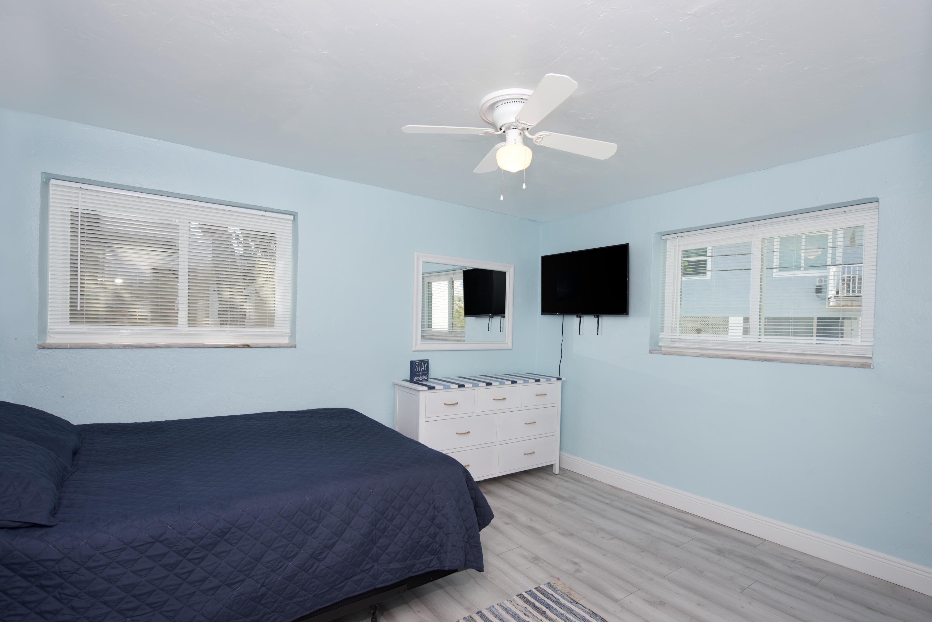 A bedroom with a bed, a ceiling fan, a dresser, a TV mounted on the wall, and windows with blinds.