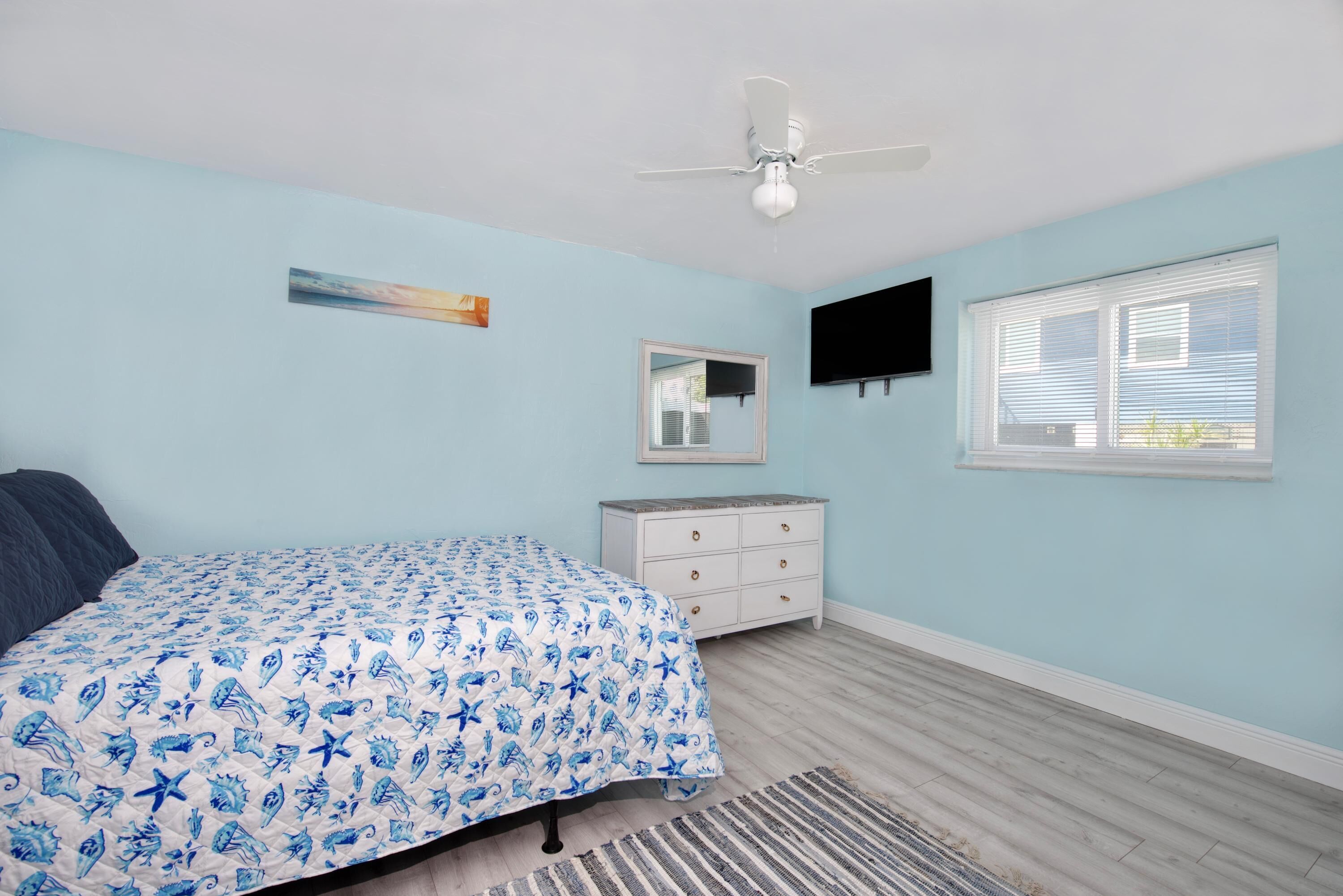 Ein Schlafzimmer mit einem Bett, einem Schminktisch, einem Deckenventilator, einem Fernseher und einem Fenster.