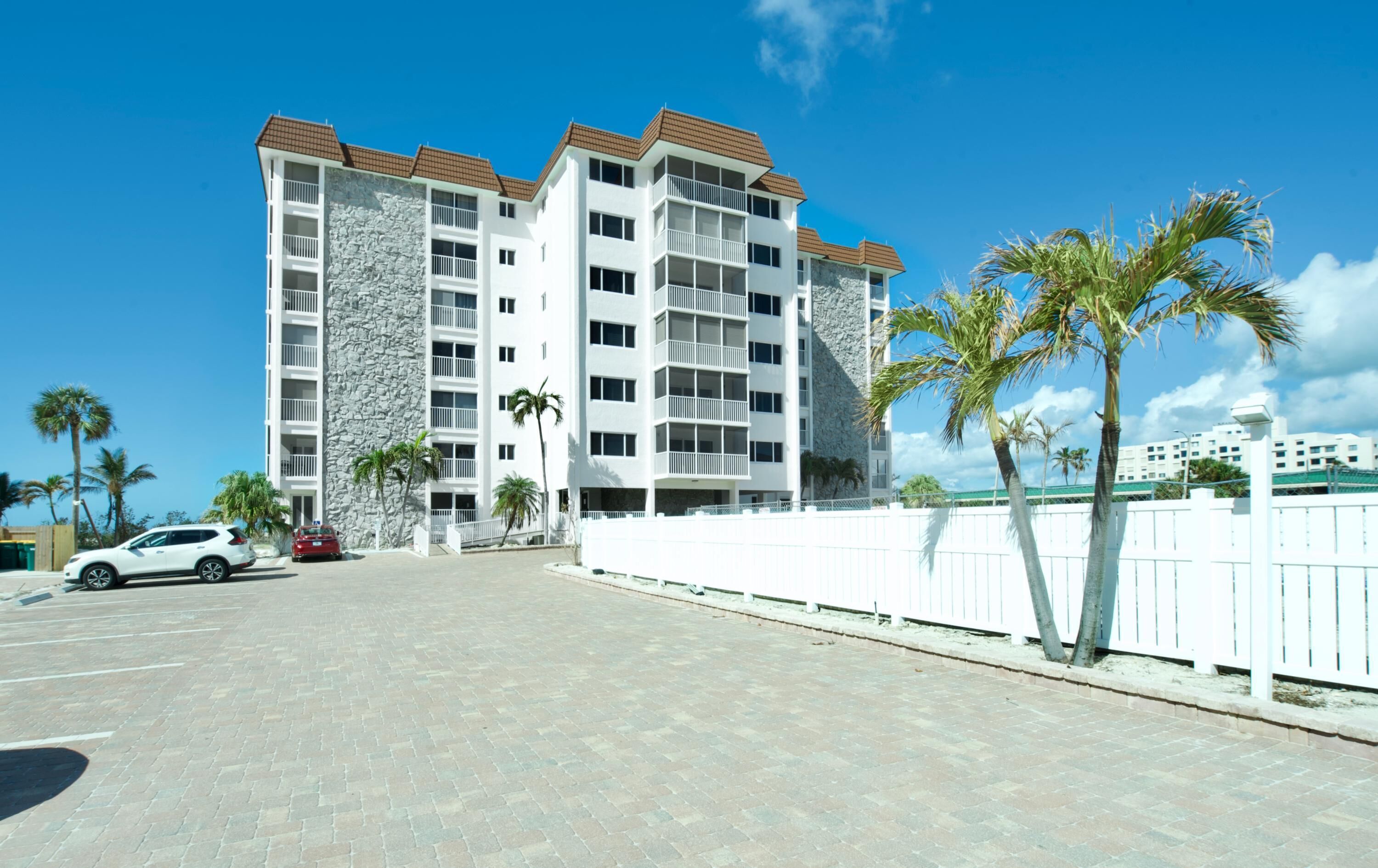 A multi-story hotel with a stone facade and balconies, a parking area, and palm trees.