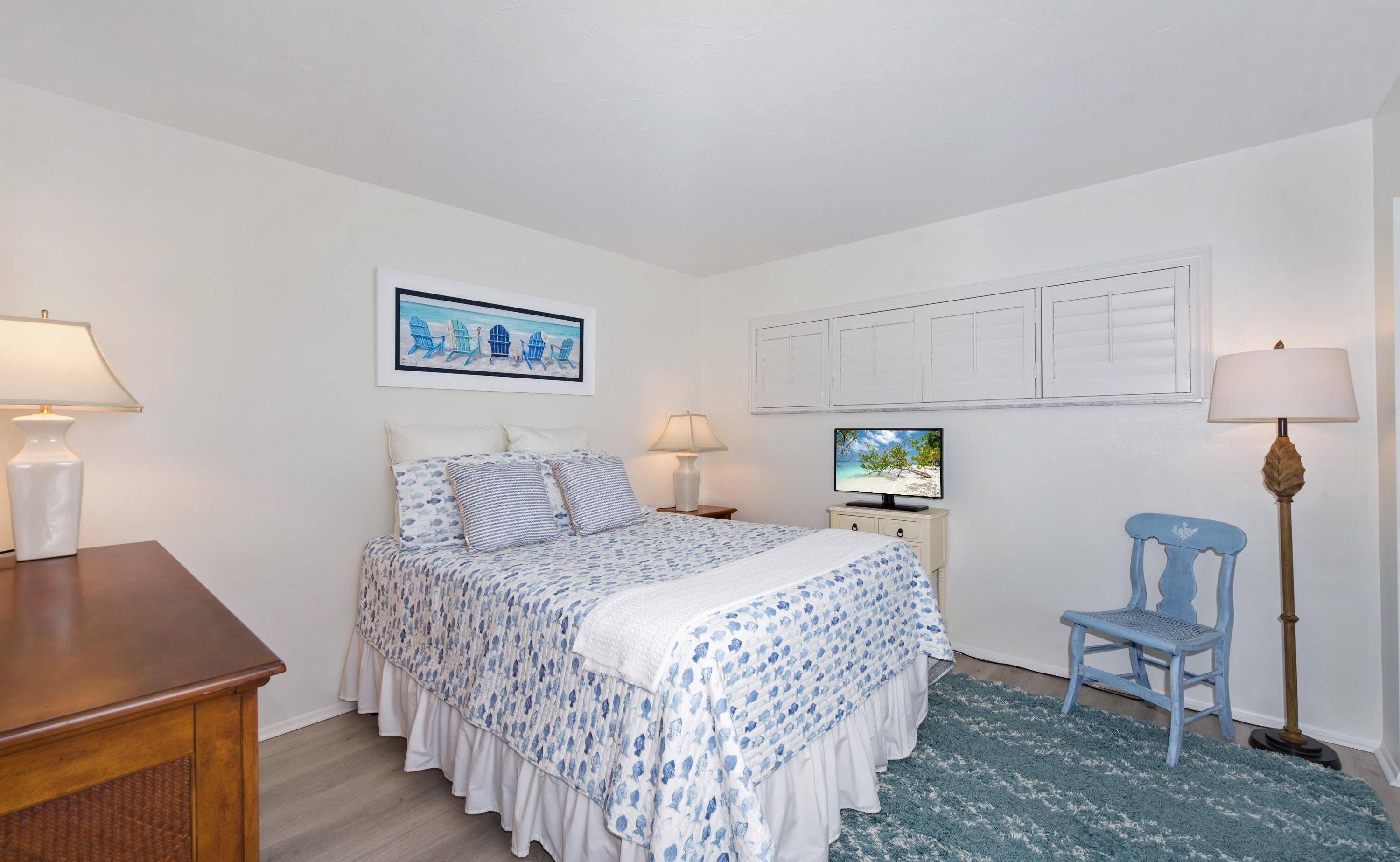 A bedroom with a bed, two bedside tables, a desk, a chair, a television, and a framed picture on the wall.