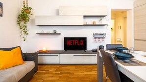 A modern living room with a sofa, a dining table with chairs, a TV displaying 'NETFLIX', and a hanging plant.