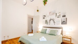 A bedroom with a bed, bedside tables, a wall with framed pictures, and a hanging plant.
