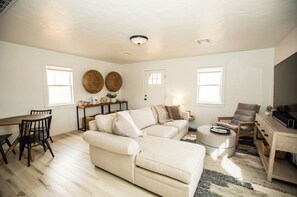 A spacious living room with a beige sofa, a wooden dining table with chairs, a small coffee table, and a TV stand. - Kanab Cottage - cozy & secluded (Kanab)