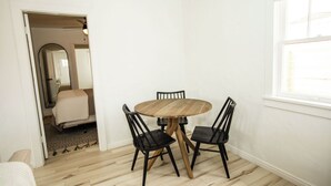 Et lille, lyst rum med et rundt træbord og fire sorte stole. Et vindue giver naturligt lys, og en dør fører til et andet rum med en seng og et spejl.