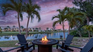 A patio with a fire pit surrounded by Adirondack chairs, overlooking a body of water and palm trees. - Endless Tides 1 (Fort Myers Beach)