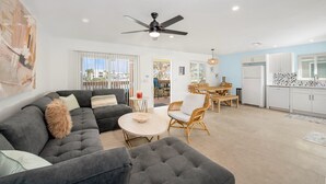A modern living room with a dark grey sectional sofa, a round coffee table, and wicker chairs. A ceiling fan is visible, and the room has large windows. - Endless Tides 2 (Fort Myers Beach)