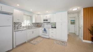A modern kitchen with white cabinetry, a microwave, a stove, and a dishwasher. There are two rugs on the floor, and the kitchen has a light blue wall. - Endless Tides 2 (Fort Myers Beach)