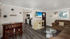 A living room with a brown couch, a glass coffee table, a wooden dining table with two chairs, a kitchen area with a microwave, and a TV on the wall.