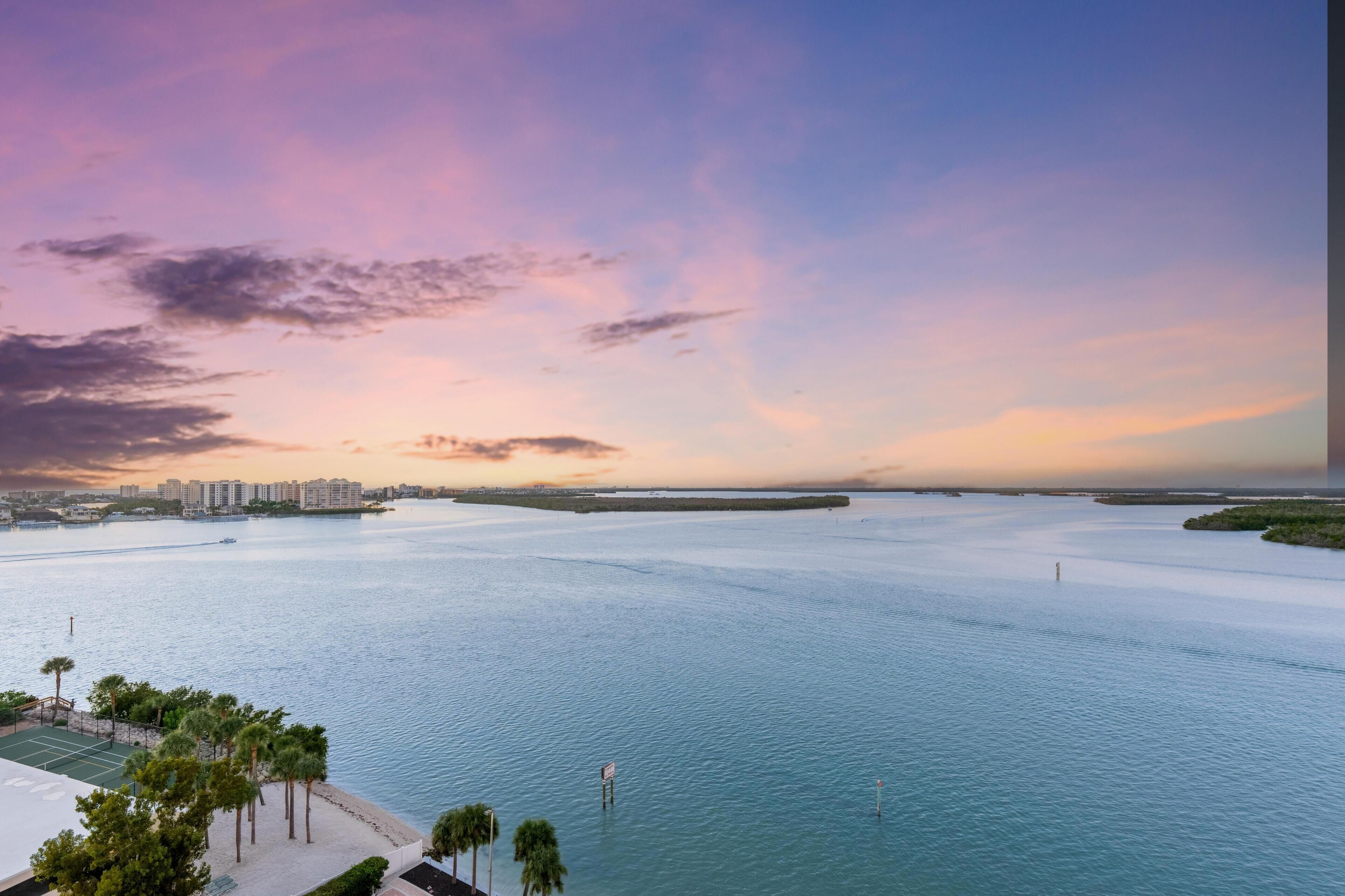 A waterfront property with a tennis court, palm trees, and a view of the city skyline.