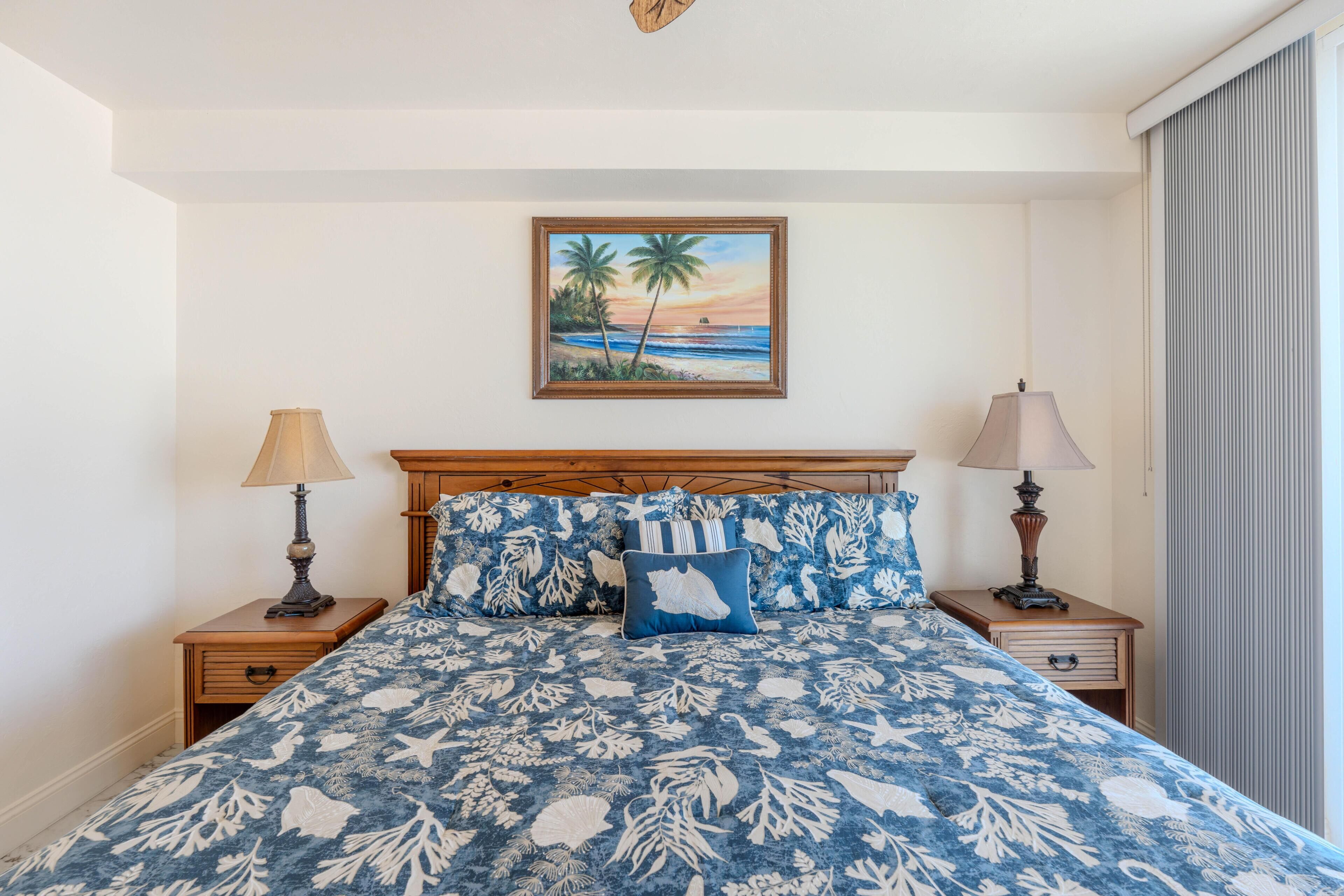 A bedroom with a bed, two bedside tables, lamps, a painting of a beach scene, and a window with curtains.