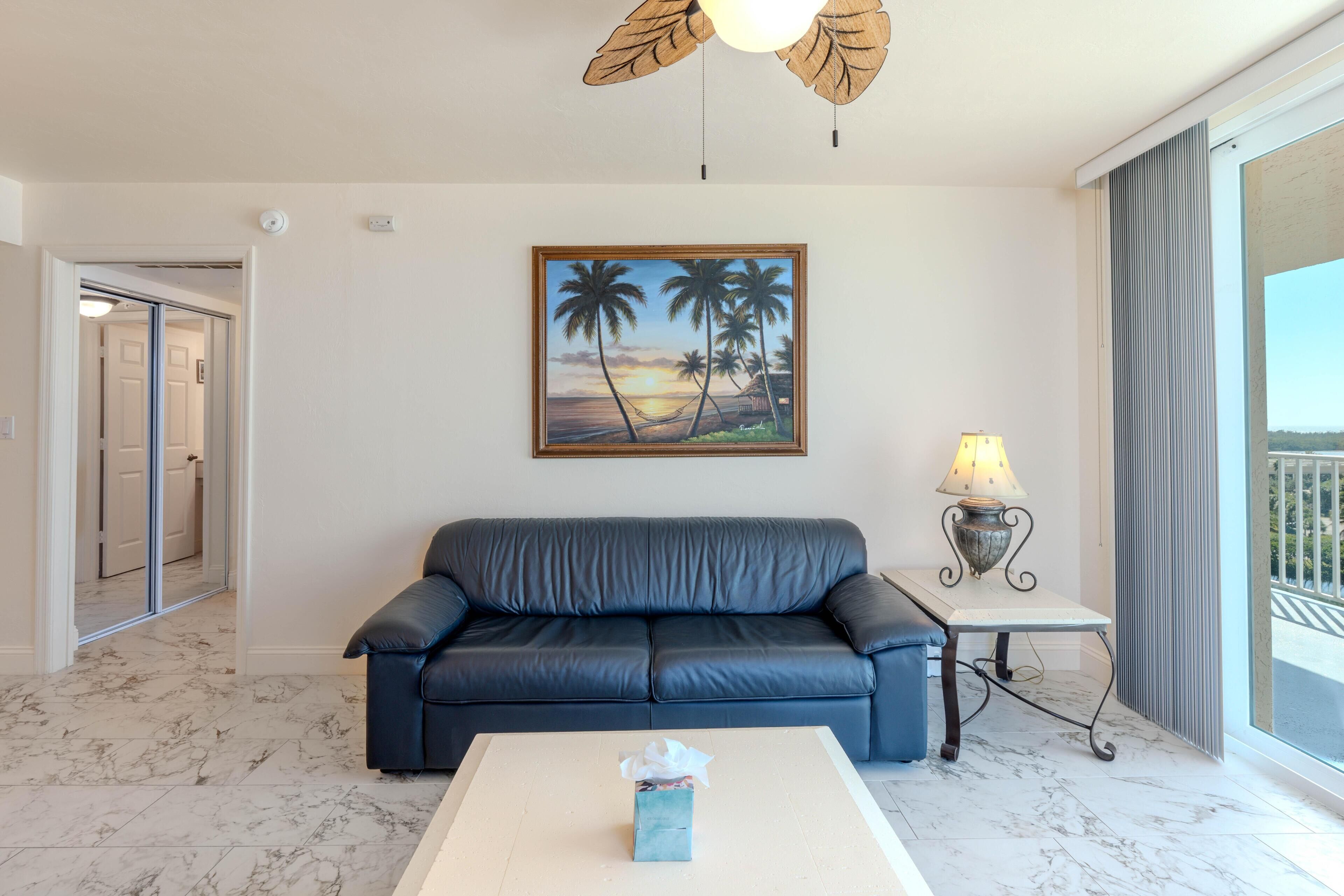 A living room with a dark blue sofa, a painting of palm trees, a lamp, and a view of the outdoors through a window.