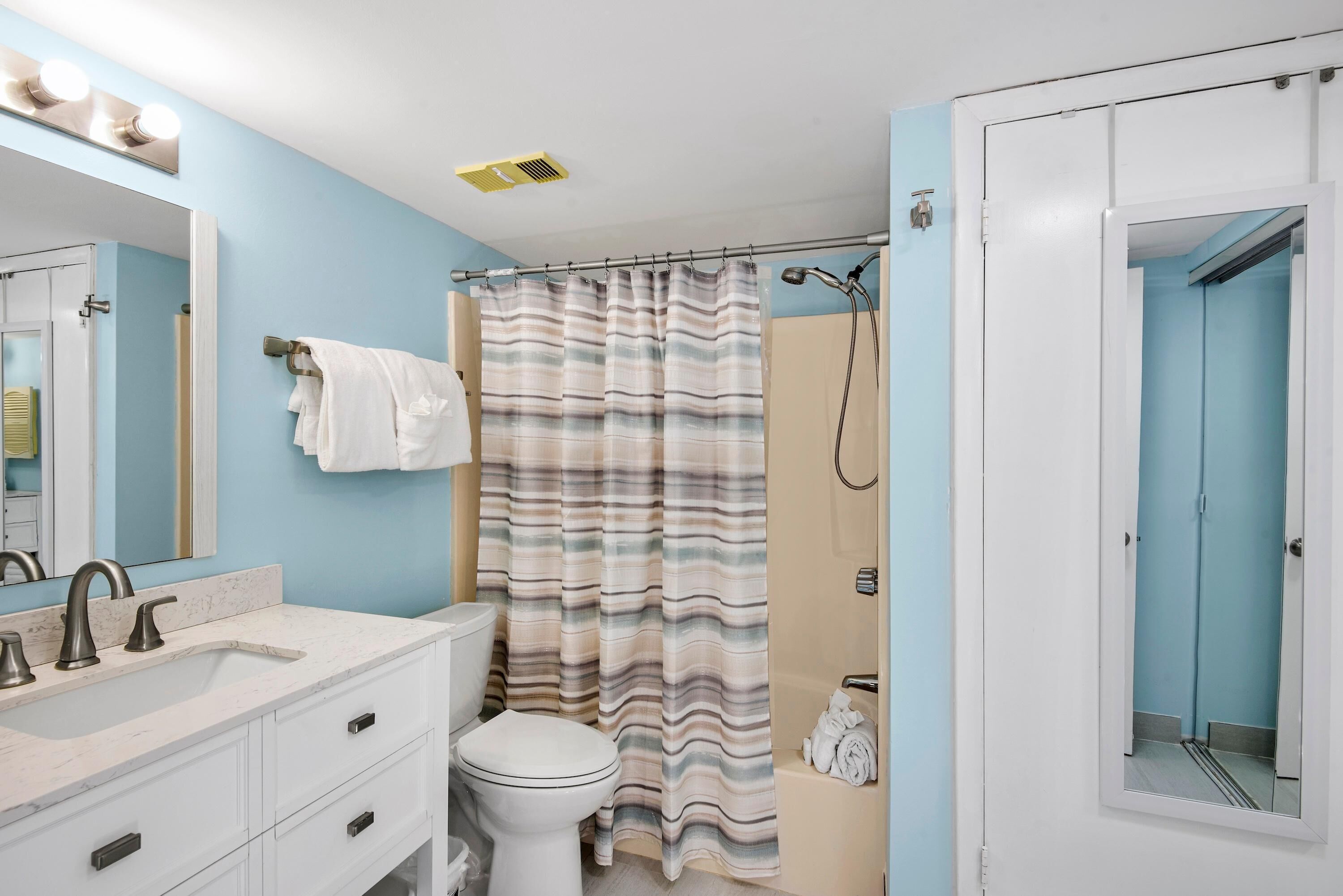 Une salle de bain avec un lavabo blanc, des toilettes, un rideau de douche à rayures et un miroir avec des lumières au-dessus.