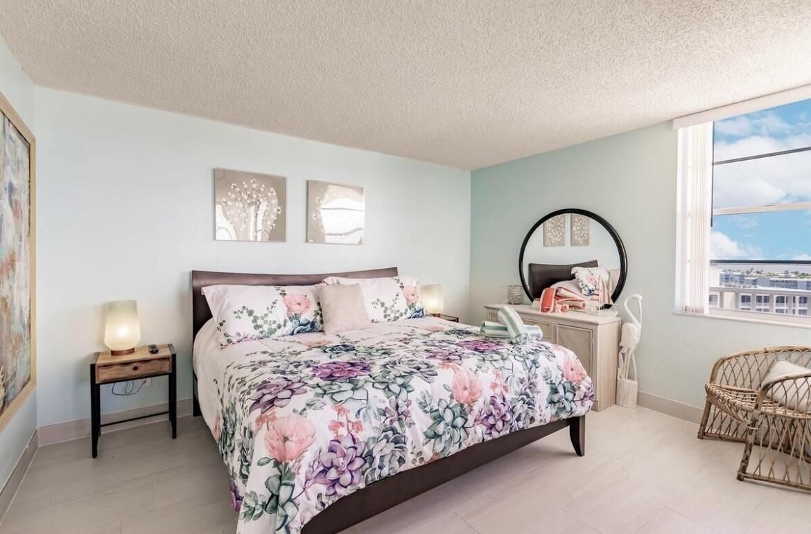 Een slaapkamer met een bloemenbedekking, een nachtkastje met een lamp, een dressoir met een spiegel en een rieten stoel.