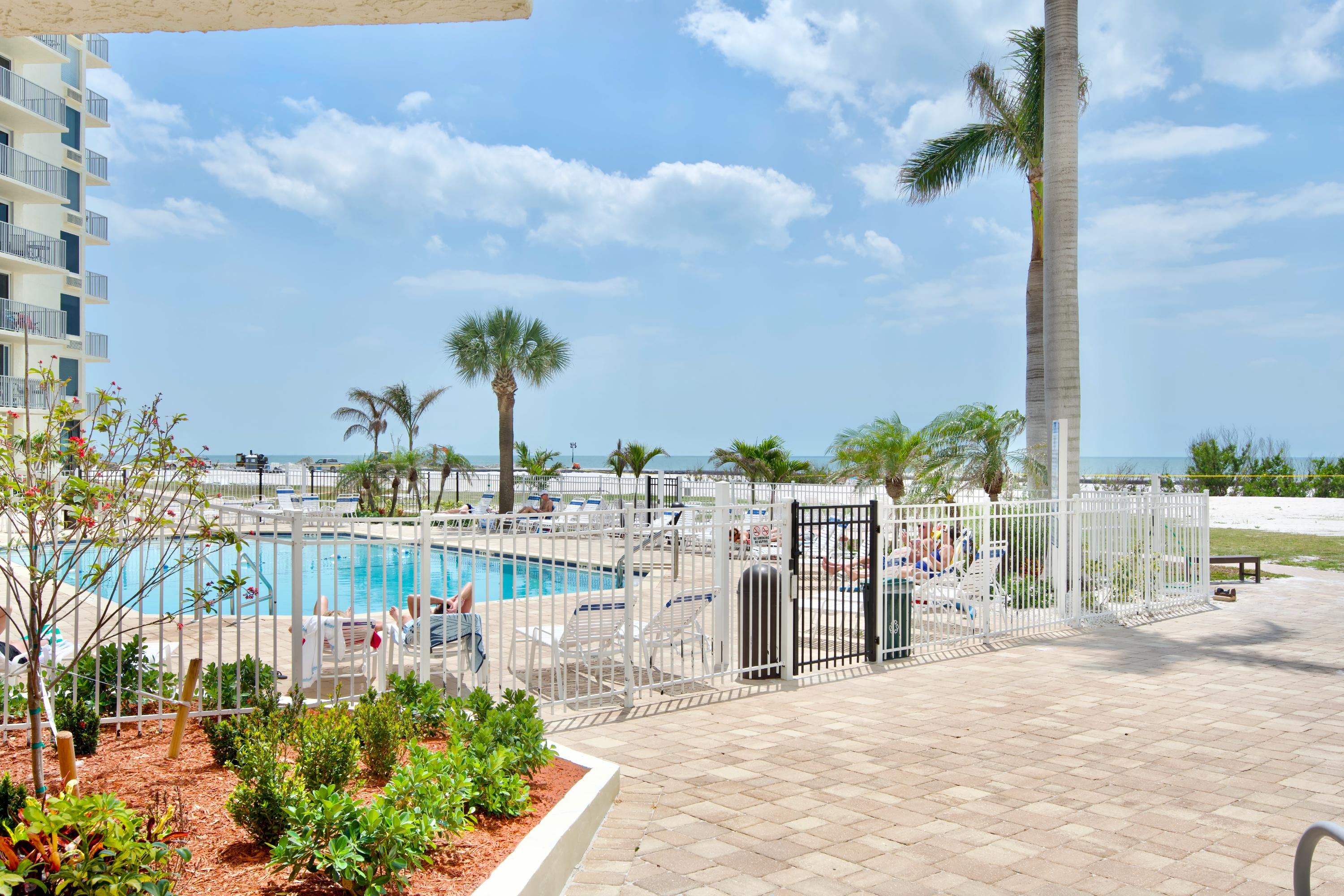 A pool area with a fenced deck, palm trees, and a view of the ocean.