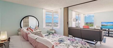 A bedroom with a bed, a desk, a chair, a TV, and a balcony with a view.