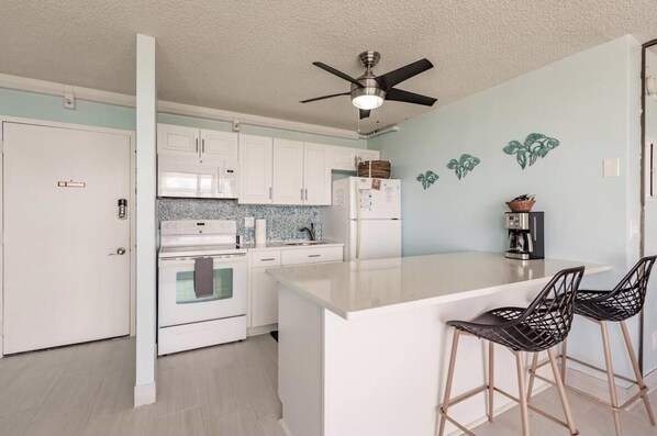 Een moderne keuken met witte kastjes, een centraal eiland, barkrukken, een plafondventilator en decoratieve wanddecoraties.