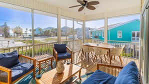 A spacious balcony with wooden furniture, a ceiling fan, and large windows offering a view of a turquoise building and palm trees. - Island Breeze (Fort Myers Beach)