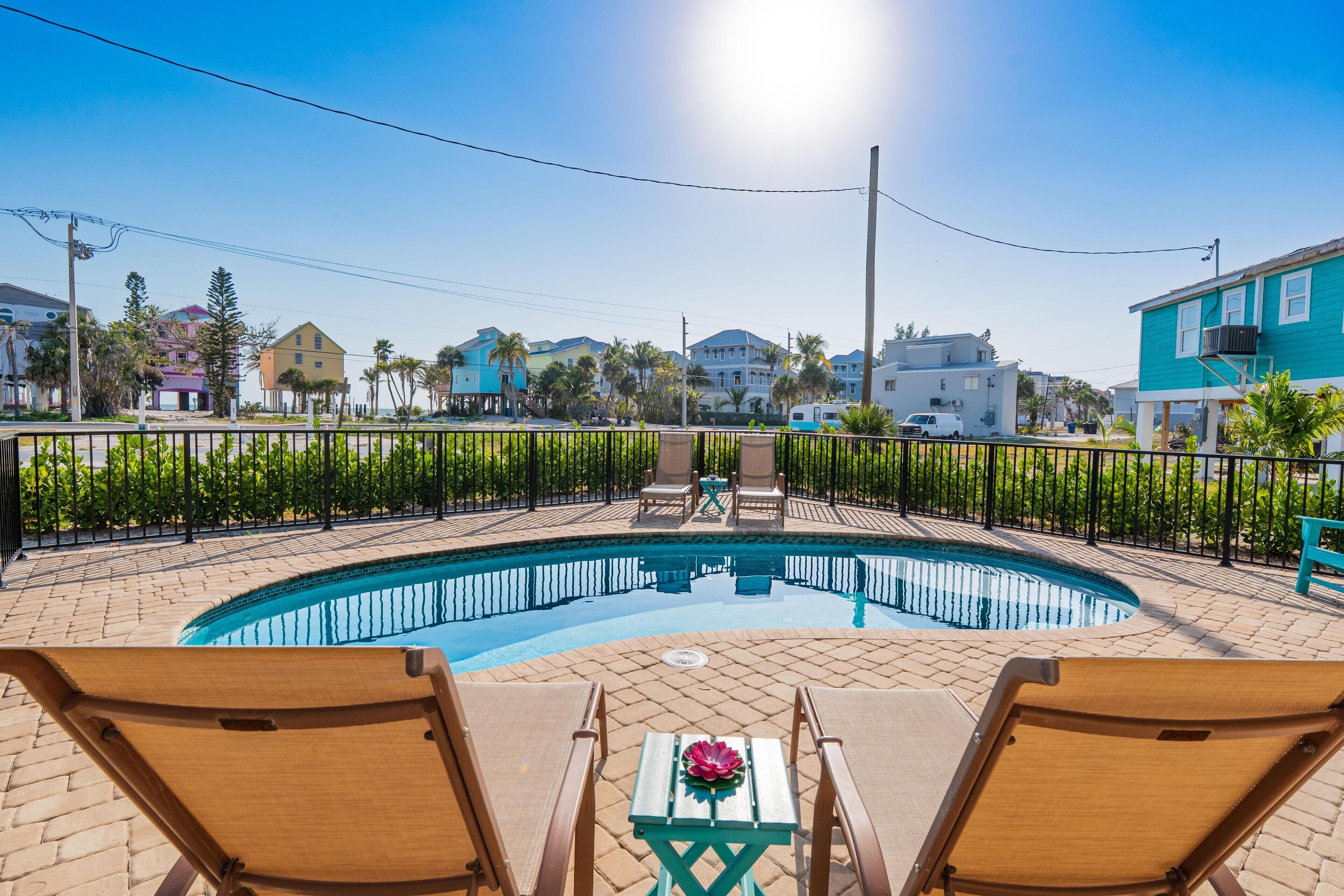 A swimming pool with lounge chairs and a table, surrounded by a fence and greenery.