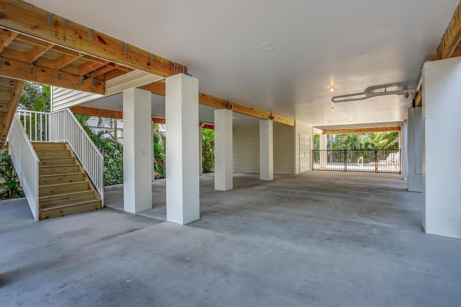 A spacious indoor area with a concrete floor, white pillars, a wooden staircase, and a view of a swimming pool outside.