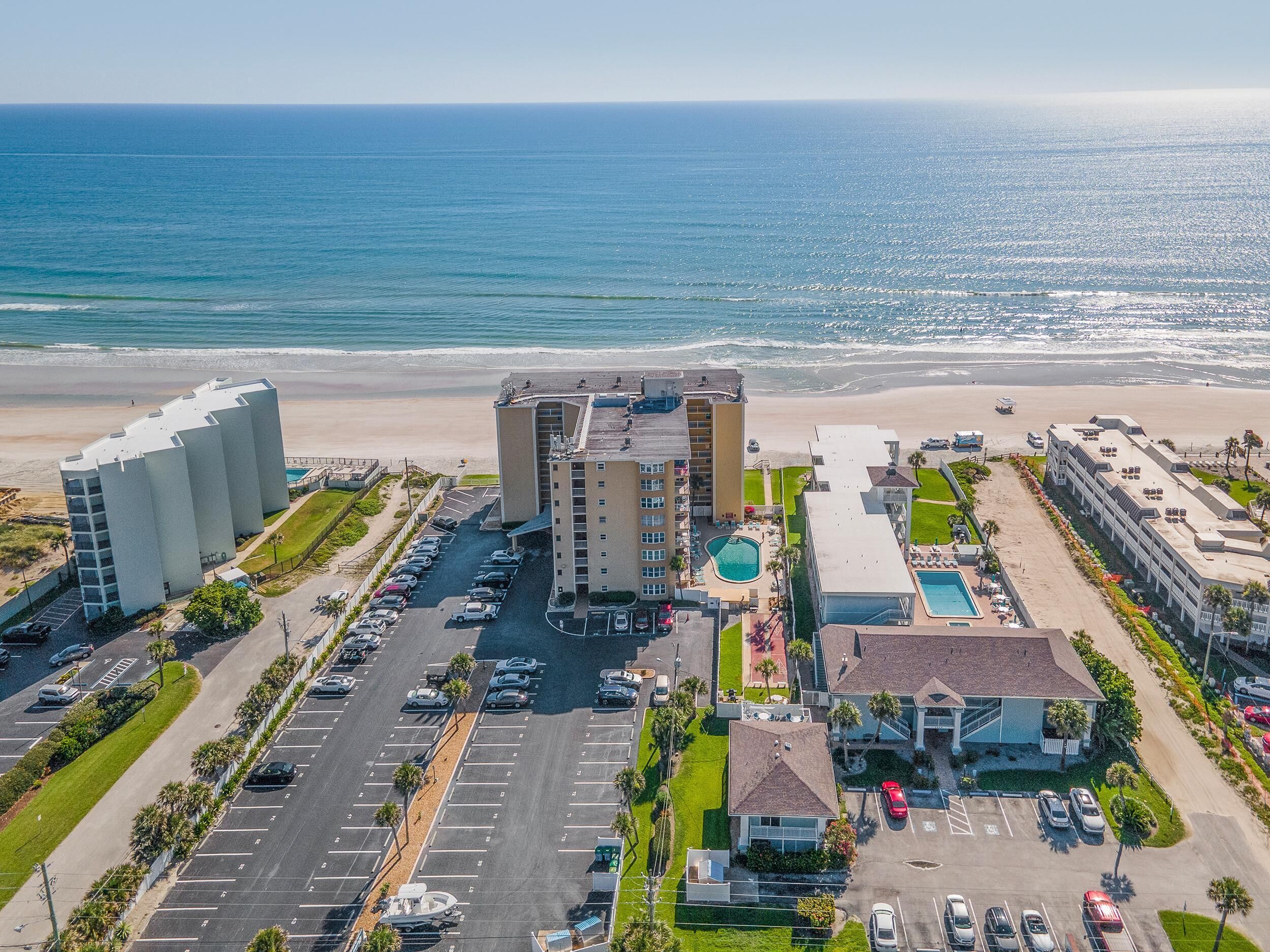 Un hotel frente a la playa con varias construcciones, estacionamiento y zona de piscina.