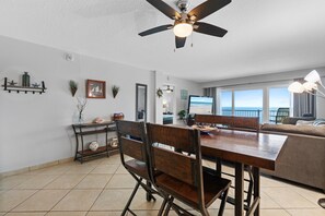 A modern living room with a dining area, a ceiling fan, and a large window offering a view of the ocean. - Ponce de Leon Towers 503 (New Smyrna Beach)