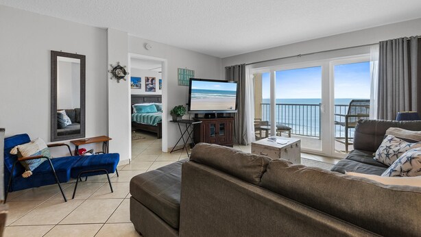 Una sala de estar moderna con un televisor grande, un sofá, un sillón azul y un balcón con vistas al mar.
