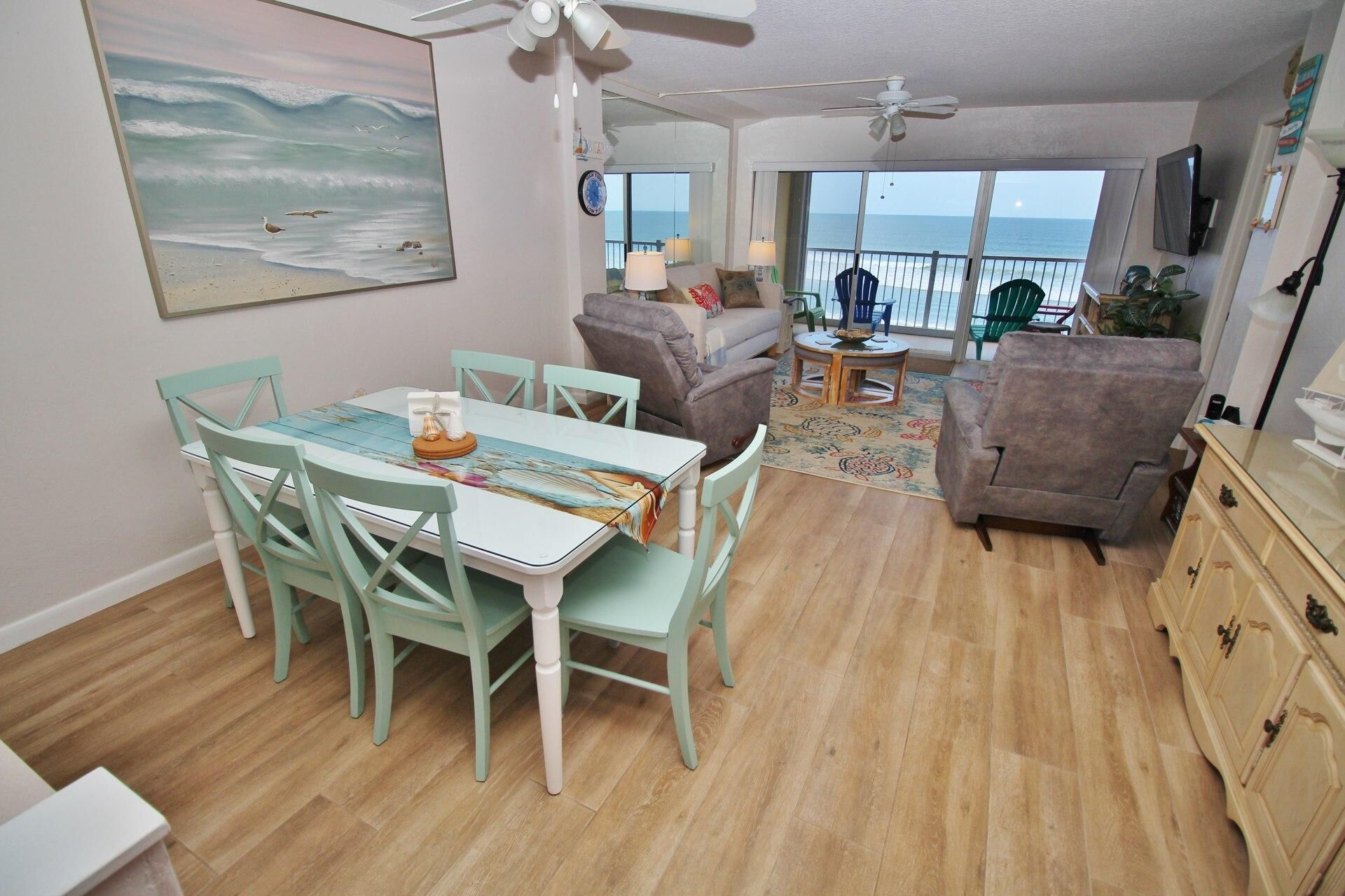 A spacious living area with a dining table, chairs, and a sofa. A large window offers a view of the ocean.
