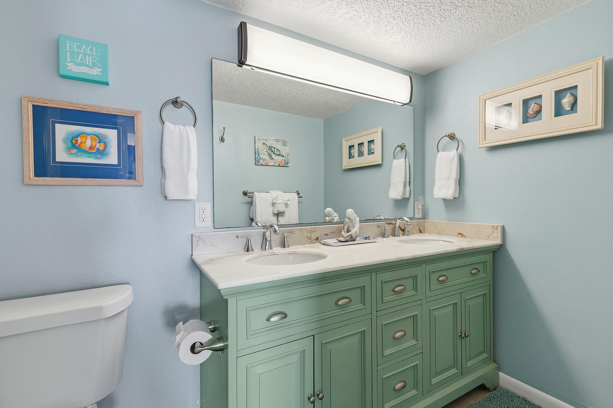 A bathroom with a light blue wall, a white toilet, a green vanity with a mirror above the sink, and framed artwork on the wall.