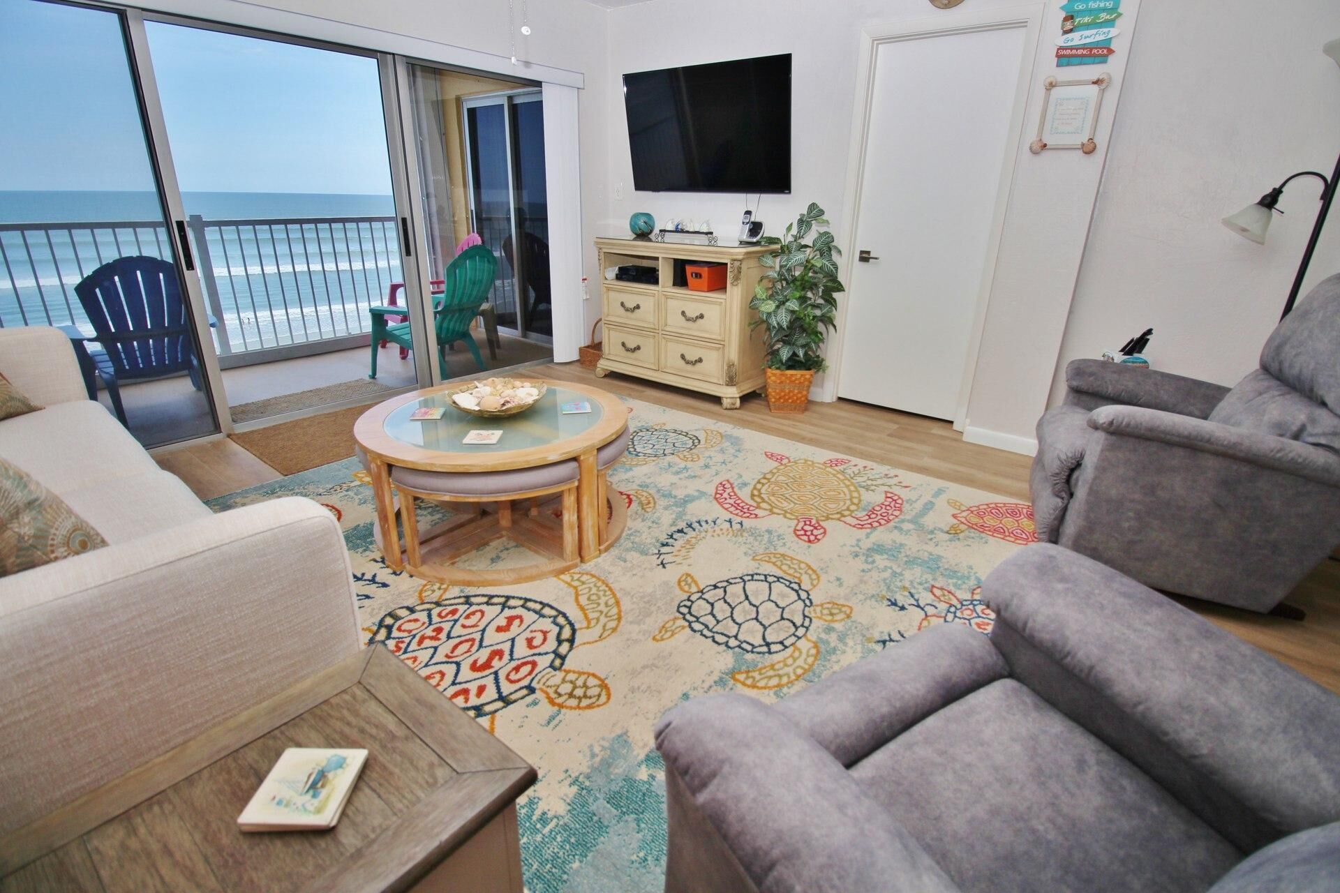 A living room with a view of the ocean, a round table with a bowl, a sofa, a chair, and a TV on the wall.