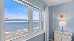 A room with a large window overlooking a beach, a white dresser with a lamp, and a framed picture on the wall. - Ponce de Leon Towers 702 (New Smyrna Beach)