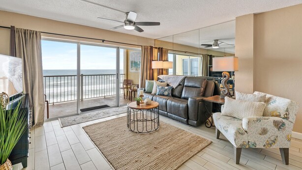 Ein geräumiges Wohnzimmer mit einem großen Fenster und Blick auf den Strand, einem Ledersofa, einem gemusterten Sessel und einem runden Couchtisch.