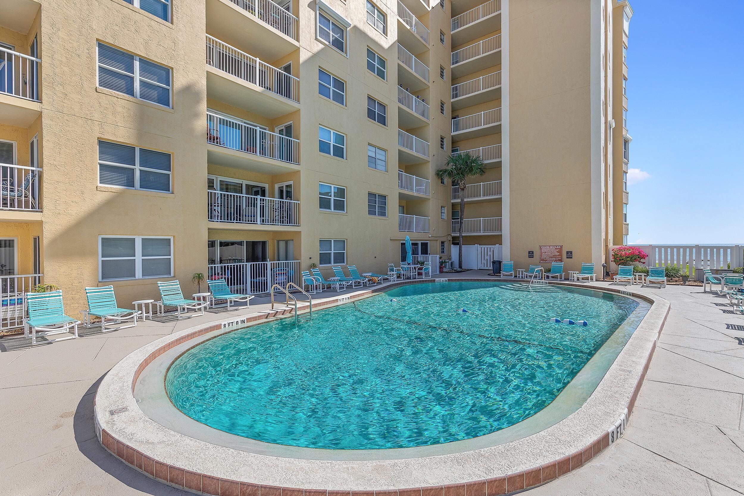 A swimming pool surrounded by a multi-story building with balconies, lounge chairs, and a clear blue sky.