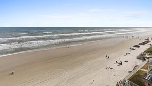 Ein Strand mit einer weiten Sandfläche, einigen Personen und einer Reihe von Liegestühlen und Sonnenschirmen.