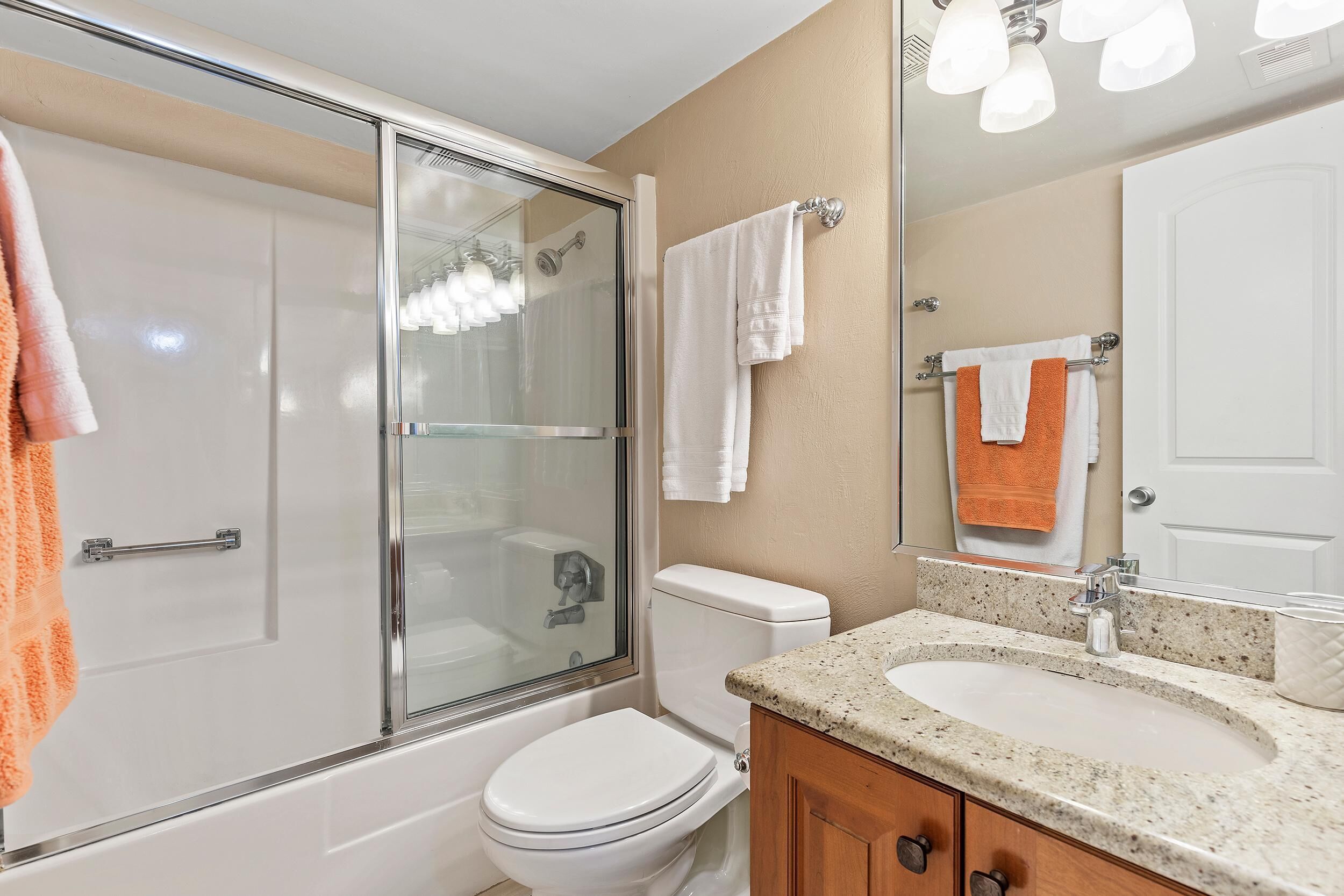 A bathroom with a glass-enclosed shower, a white toilet, a sink with a granite countertop, and a mirror with a towel rack.