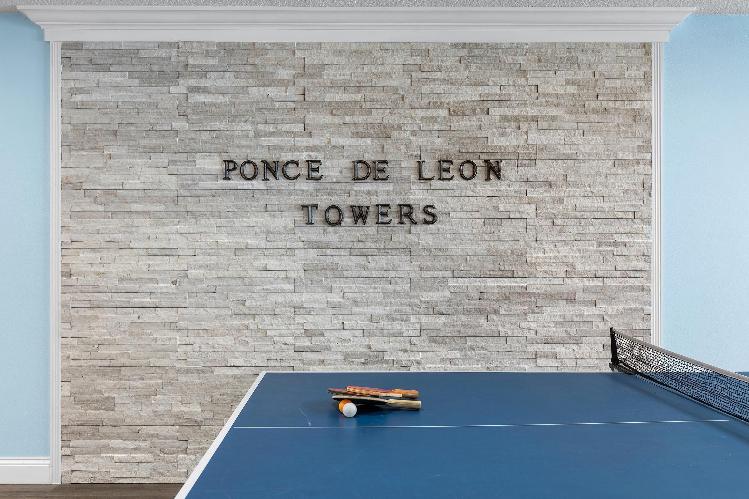 Une table de ping-pong avec des raquettes et une balle, placée contre un mur sur lequel on peut lire l'inscription « PONCE DE LEON TOWERS ».