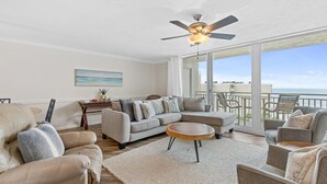 A spacious living room with a large sectional sofa, a round coffee table, and a ceiling fan. A large window offers a view of the ocean. - Pelican C606 (New Smyrna Beach)