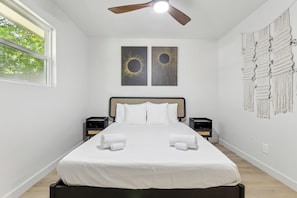 Un dormitorio moderno con una cama grande, dos obras de arte colgadas en la pared, un ventilador de techo y un tapiz de macramé.