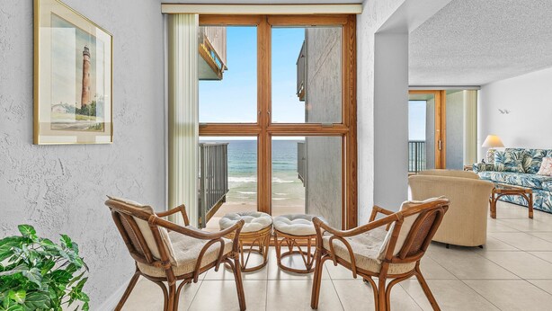 Una sala de estar moderna con un amplio ventanal que da a un balcón con vistas al mar, mobiliario de mimbre y un cuadro en la pared.