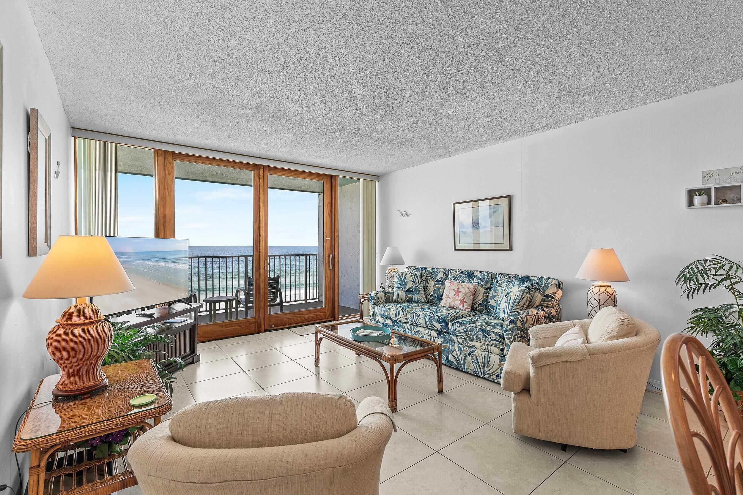 A living room with a sofa, armchairs, a coffee table, and a TV. There is a balcony with a view of the sea.