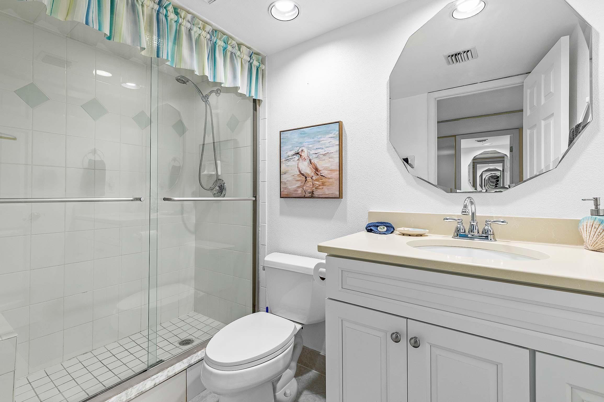 A bathroom with a glass-enclosed shower, a toilet, a sink with a mirror above it, and a framed picture on the wall.