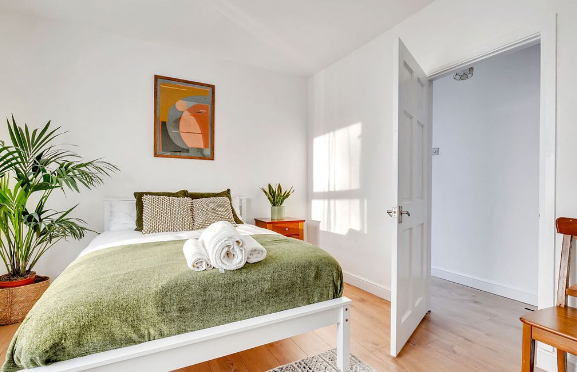 A neatly arranged bedroom with a bed, pillows, a green blanket, and a rolled towel. There are potted plants, a painting on the wall, and a door leading to another room.