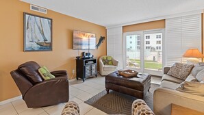 A living room with a brown leather sofa, a grey sofa, and a patterned armchair. There is a black entertainment unit, a window with blinds, and a view of buildings outside.