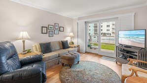 Ein Wohnzimmer mit einer Ledersofa, einem kleinen Hocker, einem Fernseher und Blick auf das Meer durch eine Glastür.