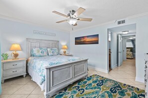 Une chambre à coucher comprenant un lit, des tables de chevet, un ventilateur de plafond, un tableau et un couloir menant à une autre pièce.