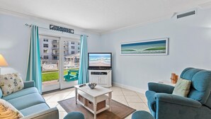 A living room with a TV, a sofa, a chair, and a view of a building with balconies. - Sea Coast Gardens II 110 (New Smyrna Beach)