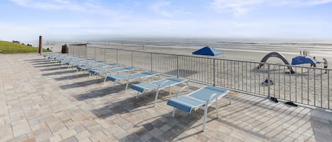 En strandnära lounge med solstolar och parasoller, ett metallräcke och utsikt över stranden och havet.