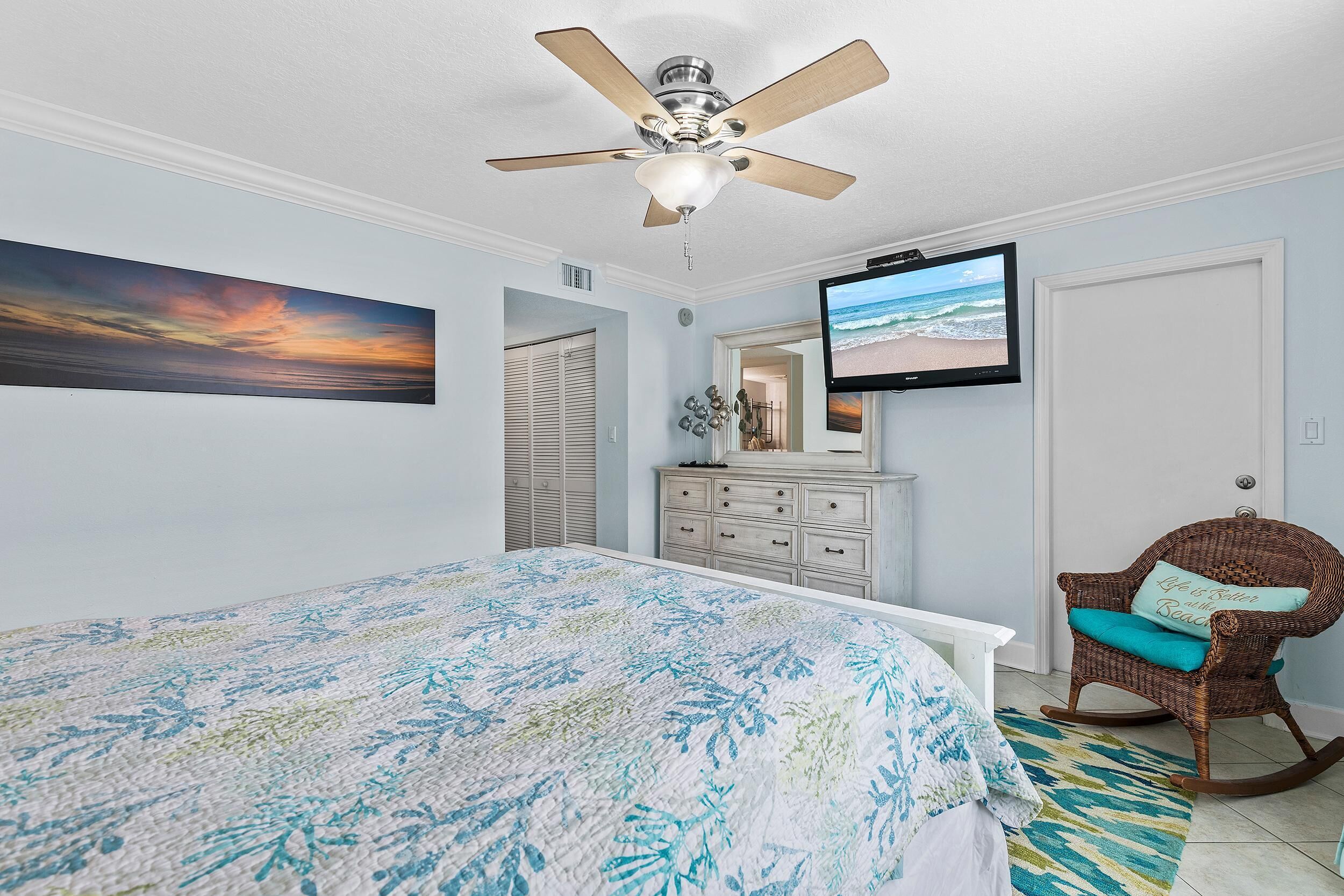 A bedroom with a bed, a ceiling fan, a TV, a rocking chair, and a painting of a sunset.